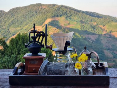 Ahşap bir masada kahve damlatan seti ve bulanık bir dağ arkaplanı Manuel kahve öğütücüsü, çaydanlığın üzerine dökülen su, kahve kurutma makinesi, 2 tadım kupası, kahve filtresi...