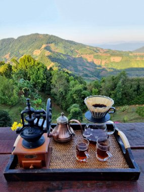 Ahşap bir masada kahve damlatan seti ve bulanık bir dağ arkaplanı Manuel kahve öğütücüsü, çaydanlığın üzerine dökülen su, kahve kurutma makinesi, 2 tadım kupası, kahve filtresi...