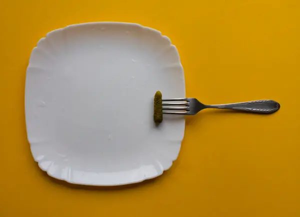 Yeşil ve küçük salatalık turşusu beyaz tabakta çelik çatal ve mutfakta sarı masa. Ev yapımı yiyecekler, koruma, sebzeler, sosyal yardımlar, sağlık. Kafeler, restoranlar, yemek şirketleri, şef