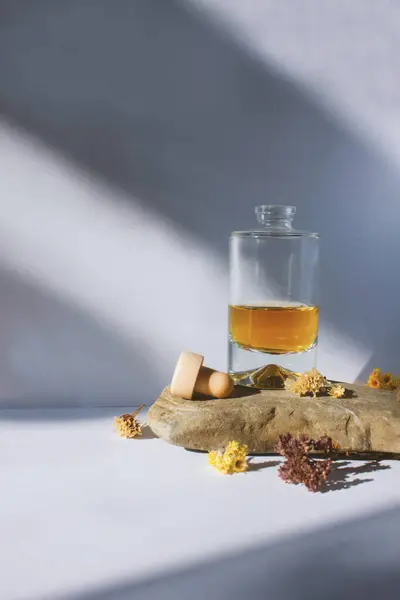 Sarı-kahverengi sıvı parfümlü berrak cam şişe, açık mantar ve kurumuş çiçekler, denizden gelen gri taşın üzerinde duruyor ve beyaz arka planda güneş parlıyor. Güzellik ve bakım, güzellik salonu..