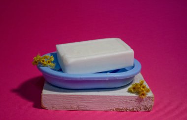 Beyaz sabun, sarı çiçeklerin yanında. Beyaz tahta bir tahtanın üzerinde mavi bir sabun kabında. Hepsi pembe arka planda. Hijyen, temizlik, sağlık. Kişisel bakım. Yüz ve vücut. Bir şeyler yıkıyorum.