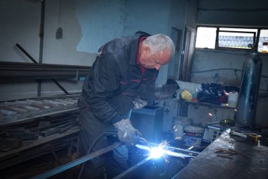 İşçi, kaynakçı, iş kıyafetleri, inşaat eldivenleri ve bir kaynak maskesiyle kaynak makinesi, Kocani Makedonya yakınlarındaki atölye çalışmasında kullanılıyor.