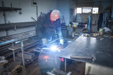 İşçi, kaynakçı, iş kıyafetleri, inşaat eldivenleri ve bir kaynak maskesiyle kaynak makinesi, Kocani Makedonya yakınlarındaki atölye çalışmasında kullanılıyor.