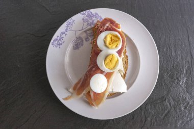 Beyaz tabakta jambonlu, peynirli ve yumurtalı sandviç.