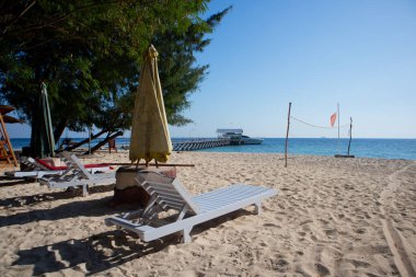 Güneşli bir günde kumlu bir sahilde bir sıra plaj sandalyesi ve şemsiye. Sandalyeler ve şemsiyelerin hepsi farklı renkte ama hepsi parlak ve neşeli görünüyor..