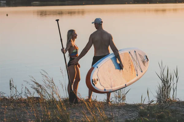 young woman with surfboard and man standing by river
