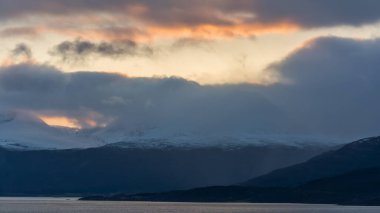 Norveç 'in kuzeyinde, gün batımında kara bulutlar ve karla kaplı fiyort kenarındaki dağlar.
