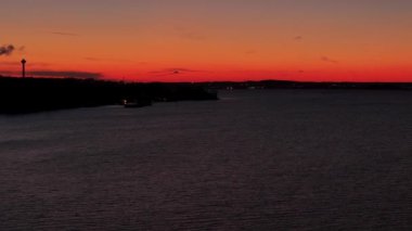 Finlandiya, Tampere 'deki binaların siluetinin ardında, karanlık orman kıyıları ve günbatımının renkleri ufukta beliren bir gölün üzerinde dönen uçuşlar.