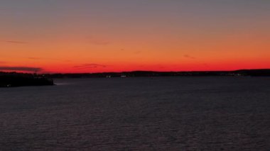 Tampere, Finlandiya 'daki binaların siluetinin arkasındaki ufukta karanlık orman kıyıları ve günbatımı renkleriyle gölün üzerinden sağa doğru yan uçuşlar.