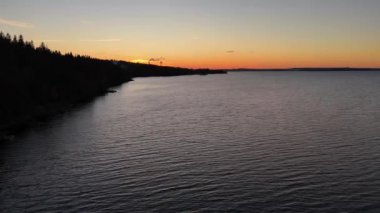 Tampere, Finlandiya 'daki binaların siluetinin arkasındaki ufukta karanlık orman kıyıları ve günbatımı renkleriyle gölün üzerinden sağa doğru yan uçuşlar.