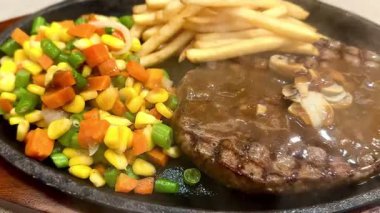 A sizzling beef steak served on a hot plate, topped with smoky barbecue sauce releases steam. The steak is paired with crispy golden fries and a side of fresh vegetatable salad.