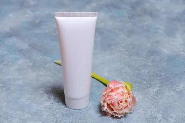White cosmetic tube and pink tulip flower on blue background. Concept minimalism in cosmetic packaging. Copy space. Natural organic spa cosmetic beauty concept.