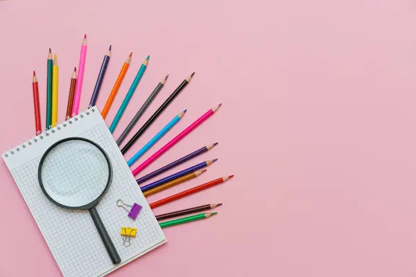 Magnifying glass, pencils and a blank white notepad on a spring on a pink background. Back to school concept. Copy space for text
