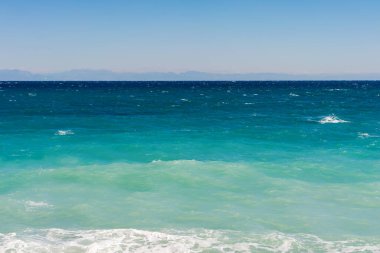 A wide view of the ocean with varying shades of blue and green waves under a clear sky. Coastal serenity and tranquility