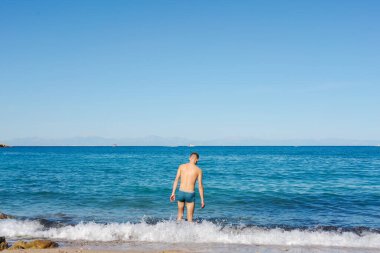 Sahilde sığ sularda duran bir adam, açık bir günde deniz ufkuna bakıyor. Rahatlama, yalnızlık ve yaz tatili kavramı.
