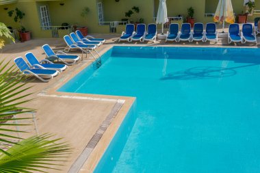 Boş havuz kenarında mavi şezlonglar ve şemsiyeler, açık mavi su, güneşli bir gün, dinlenme ve tatil konsepti.