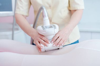 Bir kadın bir kadının bacaklarına masaj yapmak için bir cihaz kullanıyor. Cihaz beyaz ve üzerinde bir tüp var. Kadın sarı bir gömlek giyiyor.