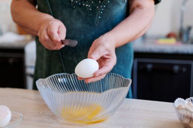 Kadın kaseye yumurta kırıyor. O, yeşil bir gömlek giyiyor. Kase ahşap bir masanın üzerinde.