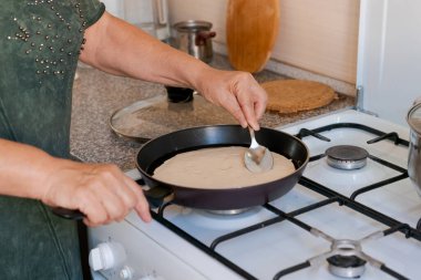 Kadın fırında krep pişiriyor. Krep hamurunu tavaya atmak için kaşık kullanıyor.