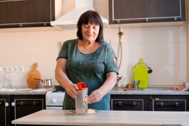 Kadın mutfakta, yemek hazırlıyor. Peynir rendelemek için yemek rendesi kullanıyor. Mutfak, lavabo, soba gibi çeşitli alet ve gereçlerle temiz ve iyi düzenlenmiş.