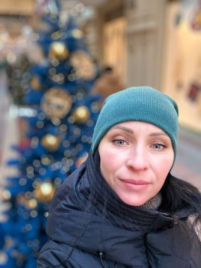 Yeşil bereli ve siyah ceketli bir kadın. Bir Noel ağacının önünde duruyor.