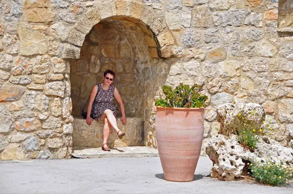 Zayıf elbiseli bir turist taştan bir duvarda, tarihi bir duvarda oturuyor ve gülümsüyor. Ön planda bir çömlek ve taş var. Kalihea, Rodos, Grece. 