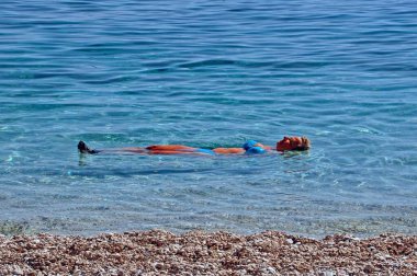 Mavi, iki parçalı mayolu bir kadın denizin çakıllı kenarında sırtüstü yatarak suda yüzüyor.. 