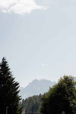 Yukarıdaki açık mavi gökyüzü ile bir dağ sırası uzaklıktan görülebilir. Ağaçlar yeşil, gökyüzü parlak ve güneşli.