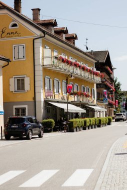 Balkonu ve kırmızı çiçekleri olan sarı bir bina. Binanın önünde bir araba duruyor.