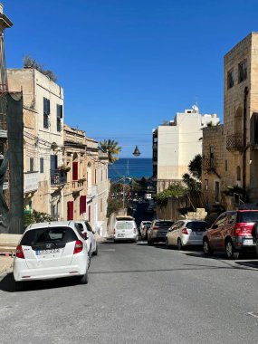 Charming Sliema caddesi, parlak binalar, mavi gökyüzü, ve park edilmiş araba.