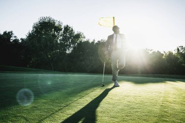Şık, kel, sakallı, şık bir takım elbiseli, kravat ve güneş gözlüklü bir zenci gün batımında özel bir golf sahasının çayırında dikilirken kulübüne ve bayrak direğine yaslanıyor.