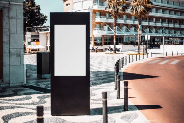 Şehir caddesinde yol kıvrımının yanında dikey dar bir pano tutucu; şehir ortamlarında boş bir reklam panosu modeli; bir tatil köyünün kaldırım taşında boş bir poster şablonu