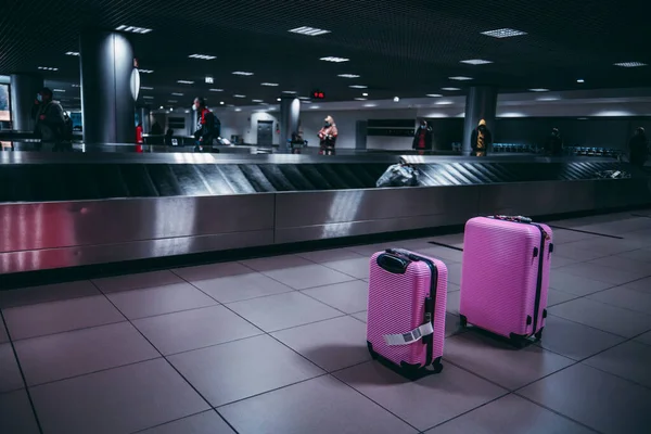 Selective focus on two pink luggage travel wheeled suitcases next to the baggage conveyor belts in an arrival area of a modern airport terminal; with a copy space area on the left for an ad message