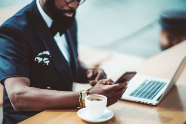 Bir sokak kafesinde sakallı bir siyahi girişimci görülüyor, elinde seçici bir odak ile akıllı telefona bir mesaj yazıyor, yanında bir fincan espresso kahvesi bir dizüstü bilgisayarın yanındaki ahşap masada.
