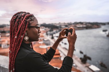 Kırmızı örgülü genç bir Afrikalı kadın yolcunun akıllı telefonuyla turistik yerleri çekerken bir profil fotoğrafı; cep telefonuyla şehir manzarasını fotoğraflayan siyah bir turist kadın.