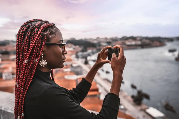 一个年轻的非洲女旅行家用她的智能手机用红色的盒子辫子拍摄旅游景点的侧面照片 一个黑人女旅行家用手机拍摄城市风景全景 — 图库照片