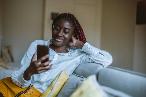 A portrait of a cheerful youthful black female sitting in her room on the grey sofa and having a video conversation via the smartphone; young black woman having a videocall using her cellphone