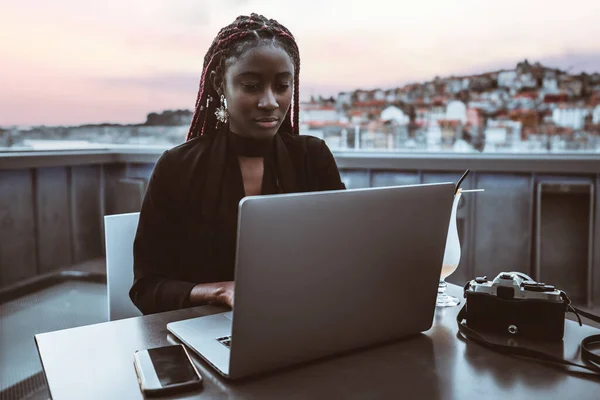 A portrait of a dazzling young black female hipster freelancer with red box braids working on her laptop while sitting on the roof cafe with a cityscape and pink sunset in a defocused background