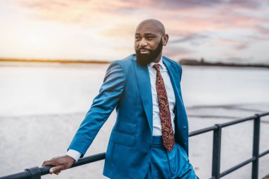 Modaya uygun, kel, sakallı, mavi takım elbiseli, kravatı çite dayanmış bir girişimci portresi. Açık arazide takım elbiseli yakışıklı bir Afrikalı işadamı.
