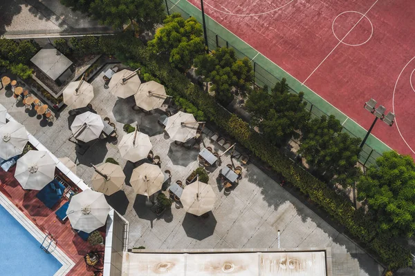 A drone view from high above of a resort restaurant with sunshade canopies, tables, and chairs next to an empty swimming pool and a basketball field on the other side separated by the tree-line