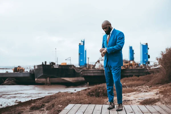 A fashionable bald mature black man is looking at his wristwatch, checking the time, while standing on a wooden pier with three metal supports in the background of the same blue color as his suit
