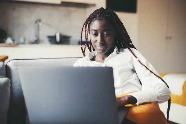 A portrait of a dazzling youthful black girl freelancer with pink color box braids working on her laptop while sitting on the coach in her living room, with a defocused background