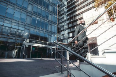 Beyaz yakalı Afrikalı bir işçinin metal bir tırabzanla merdivenlerden çıkarken çekilmiş bir fotoğrafı. Haftasonu çantası, gözlük ve çağdaş bir ofis binasından çıkan koyu renk bir takım elbise.