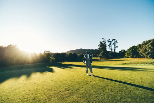 A capture of an African man, with a hipster style, posing with a golf club in his hand on a sunny day, clear sky, in a lush green golf field with shrubs and the sun flare in his back