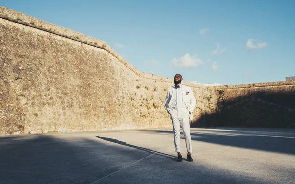 An African bald and bearded man well-dressed in a white suit with a bow tie, glasses, and leather shoes stands on the concrete floor; Surrounding him is the old wall of the village of Cascais