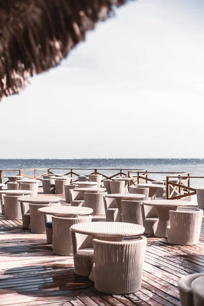 View of an empty outdoor restaurant area filled with wicker tables and chairs, bellow it a wet wooden material from the humidity and the splash of waves, in the next layer of the image is the ocean