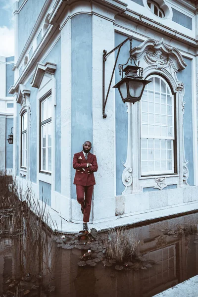 A dandy African American bald man with an unshaved beard wears a Bordeaux tailored suit while leaning against the corner of a blue edifice with classic style with a lagoon surrounding the both of them