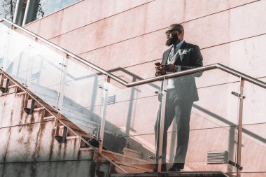 A handsome bearded African man holding a cell phone while standing on the staircase with stone damp patches on a sunny day, a leather weekender bag on the step wearing a suit and sunglasses
