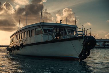 A view of a white-colored sailing boat shot at sunset. Several car tires are attached to it's rusty surface by metal clips anchored with clouds covering the sun rays, leaving a dramatic ambiance