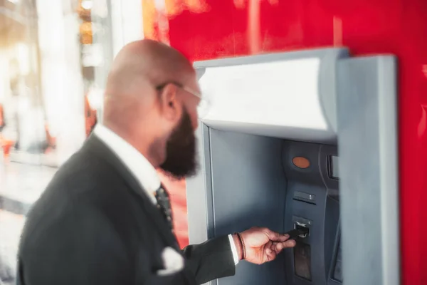A shallow depth-of-field shot with a selective focus on an elegant hairless black man\'s hand, inserting a credit card into the red and white ATM machine to withdraw cash